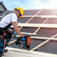 Construction worker wearing a yellow helmet and gloves using a nail gun on a wooden roof frame.