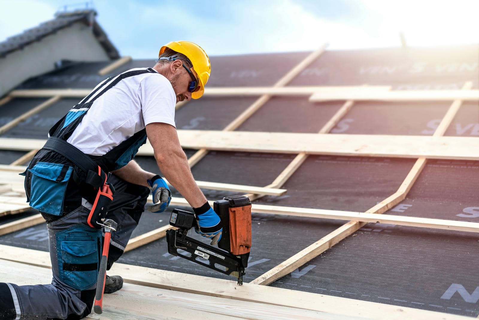Construction worker wearing a yellow helmet and gloves using a nail gun on a wooden roof frame.