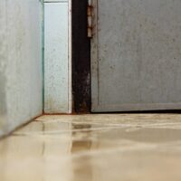 Close-up of a wet tiled floor corner next to a metal door and wall with water droplets.