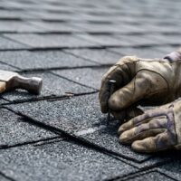 Gloved roofer hammering nails into asphalt shingles.