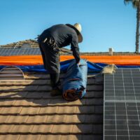 A person in a hat rolls up a blue tarp on a tiled roof next to solar panels.