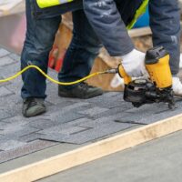 Worker using a nail gun to install asphalt shingles on a roof.